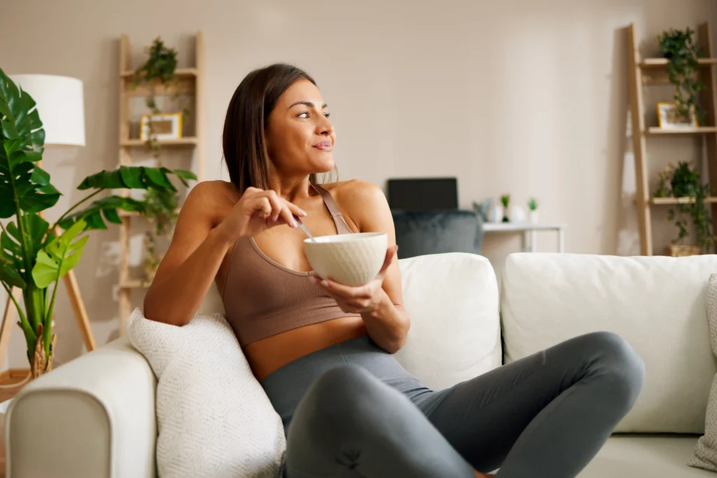 A woman in athletic wear sits on a cozy couch, enjoying a bowl of healthy food, symbolizing controlled eating habits and addressing the question: does tirzepatide suppress your appetite?