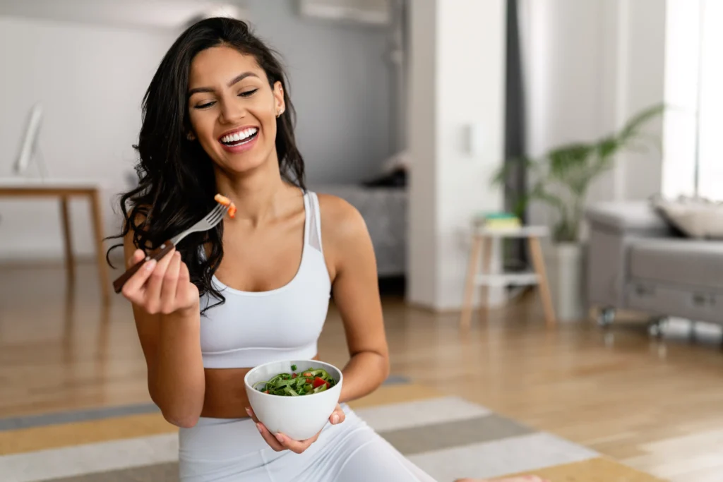 A smiling woman in athletic wear enjoys a fresh salad in a modern home setting, representing a healthy lifestyle and the benefits of a medical weight loss program.