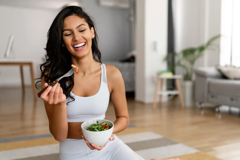 A smiling woman in athletic wear enjoys a fresh salad in a modern home setting, representing a healthy lifestyle and the benefits of a medical weight loss program.