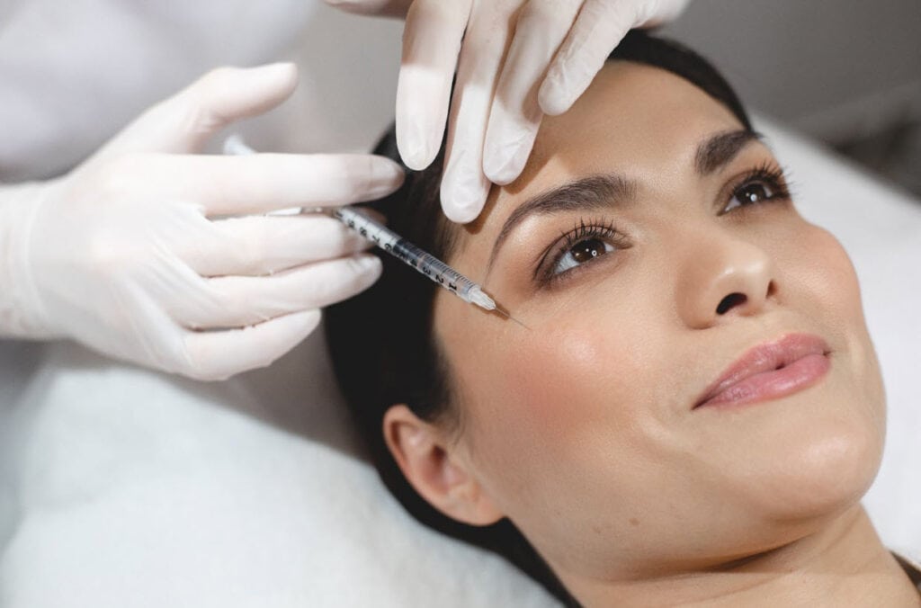 A woman with smooth skin lies on a treatment bed while a professional wearing gloves carefully administers botox under your eyes with a fine needle, targeting fine lines and wrinkles for a refreshed and youthful appearance.
