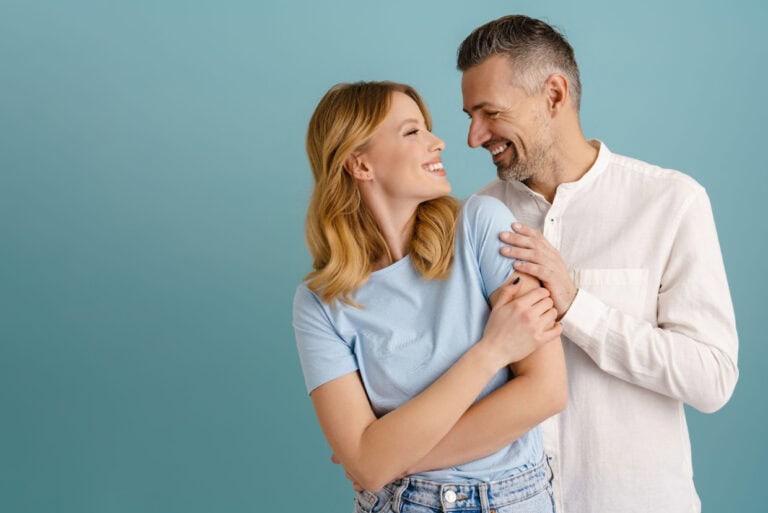 Happy middle-aged couple smiling at each other against a blue background, symbolizing improved well-being and connection after they start hormone replacement therapy.